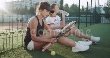 网球比赛结束后，放松时间两位年轻女士在网球场的地板上坐着，喝着水，一个